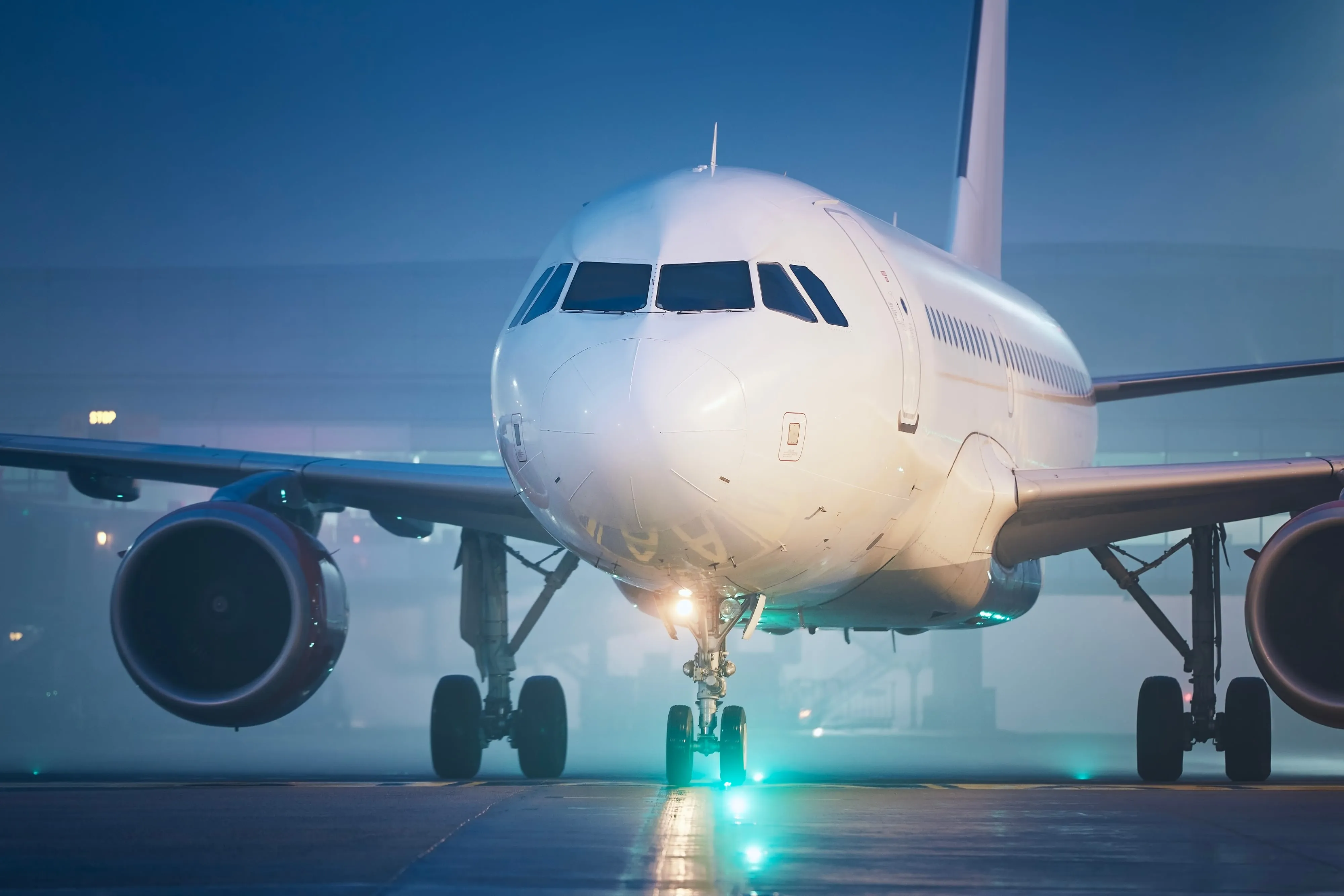 The nose of an airliner on a taxiway at dusk, lit by ground lights