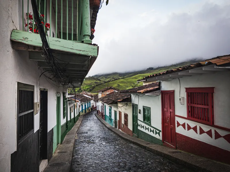 A cobbled street of whitewashed houses with brightly painted doors and window frames in an Andean coffee town, green hills rising behind