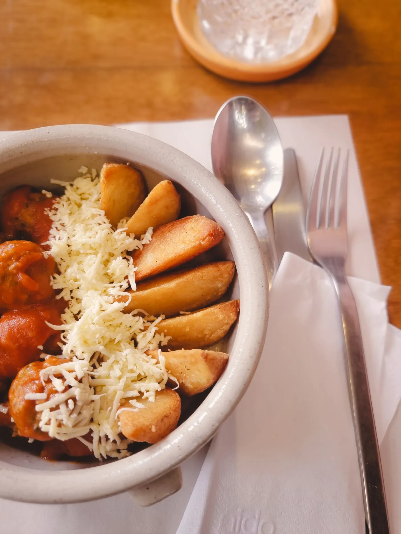 A clay bowl of stew topped with fried potato wedges and grated cheese, with cutlery alongside