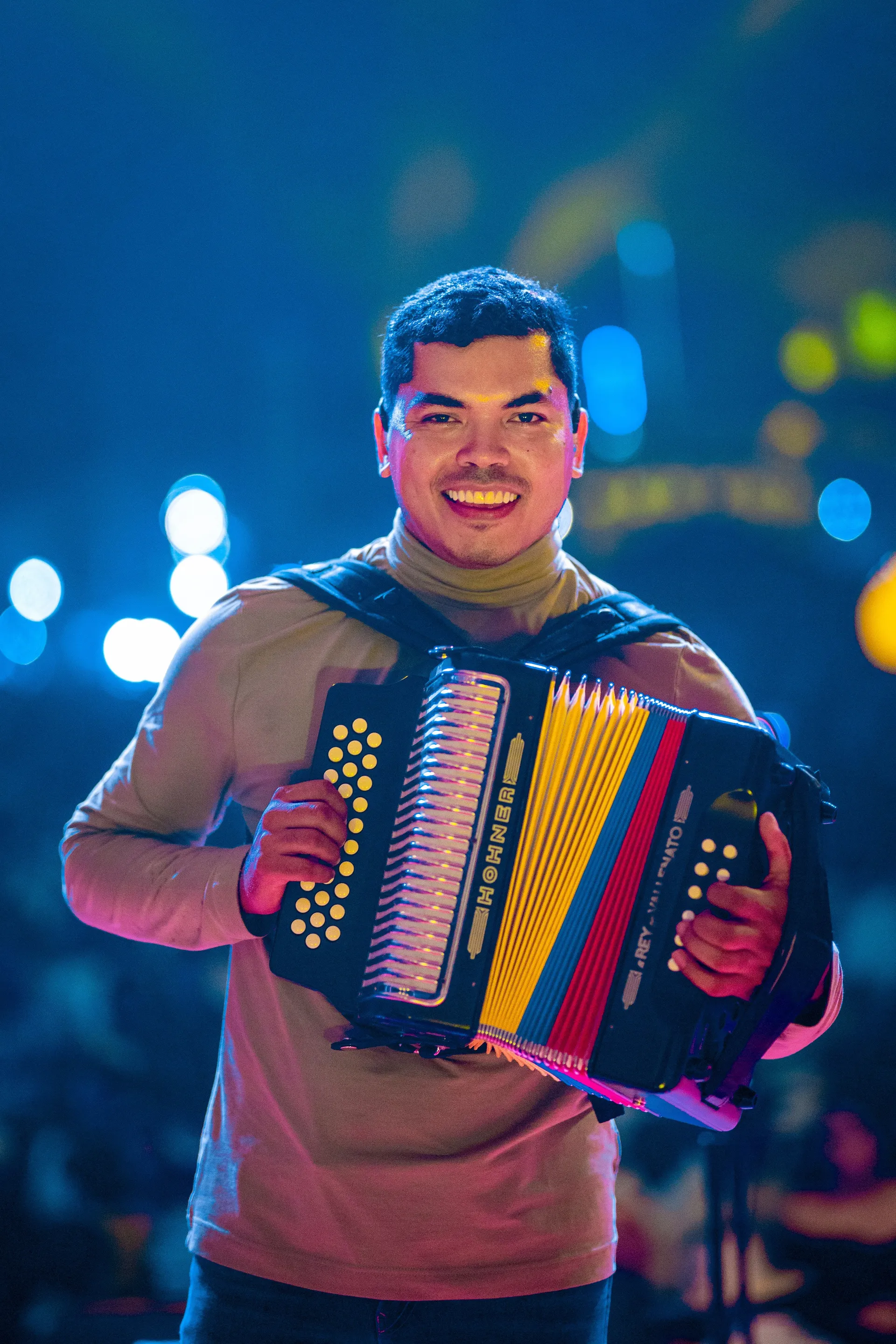 A smiling vallenato musician on stage holding a button accordion with yellow, blue, and red bellows