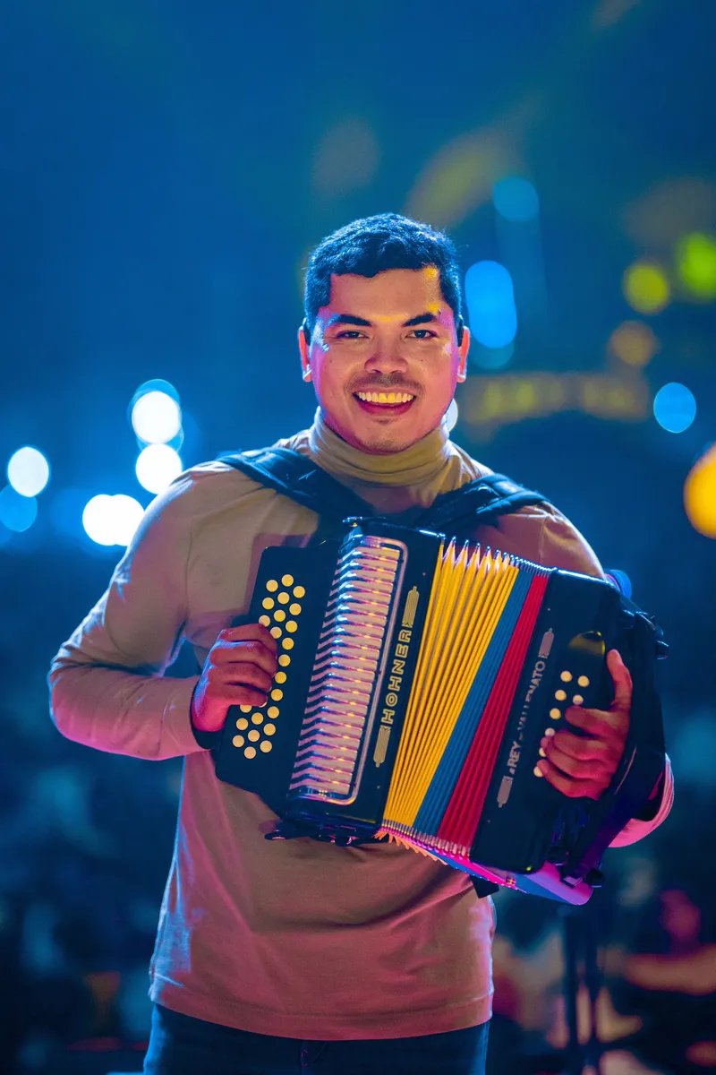 A smiling vallenato musician on stage holding a button accordion with yellow, blue, and red bellows